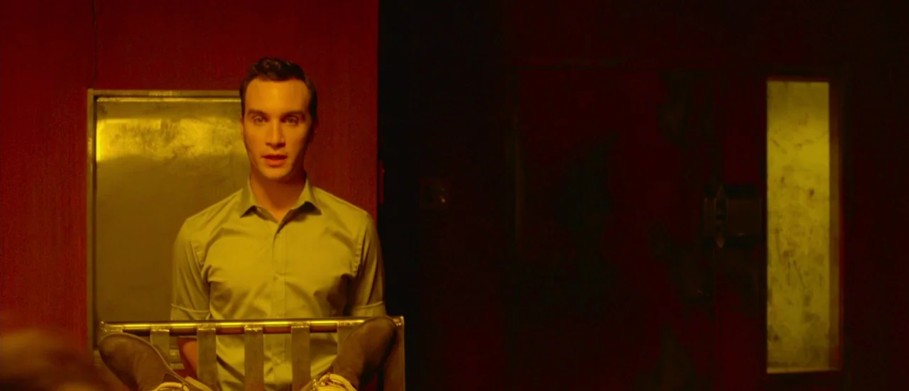 Rupture (2016) movie still: medium — A man sits behind a metal table in a dark, red-walled room. He is positioned centrally, illuminated by an…
