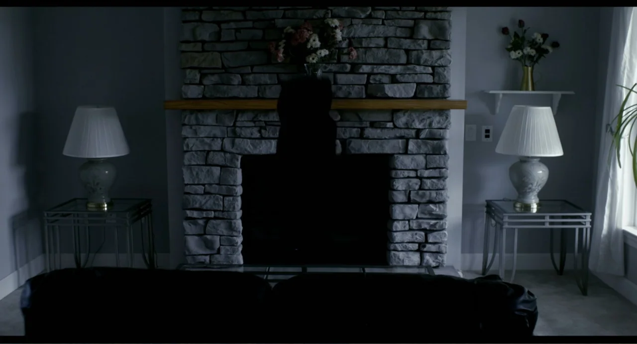 The Secret Village (2013) movie still: medium — A residential living room features a large stone fireplace with a wooden mantelpiece. A silhouetted…