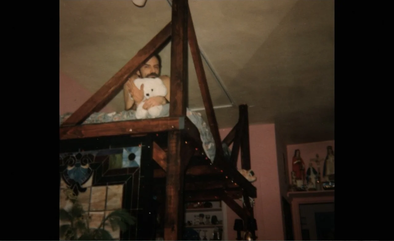 Let the Canary Sing (2023) movie still: medium — A man rests on a high wooden loft bed frame in a dark room with pink walls. Religious figurines sit…