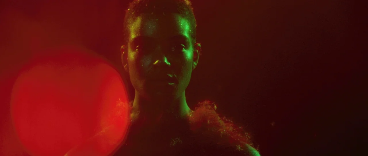 After Blue (2021) movie still: closeup — A dark space lit with intense red and green stage lighting. A person with short hair stares into the lens.