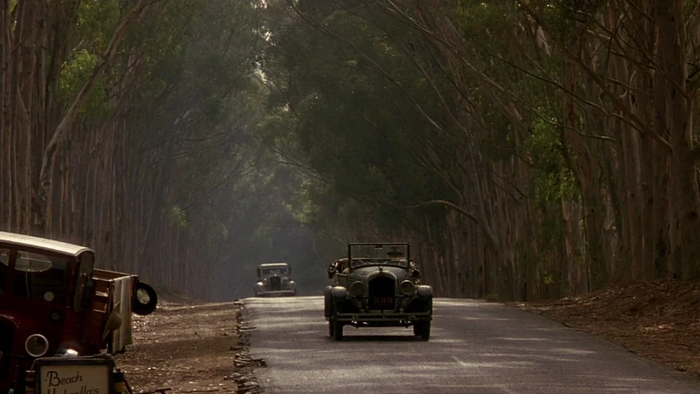 Ask The Dust (2006) movie still: wide — A narrow asphalt road winds through a dense tunnel of tall, slender eucalyptus trees. Two vintage…