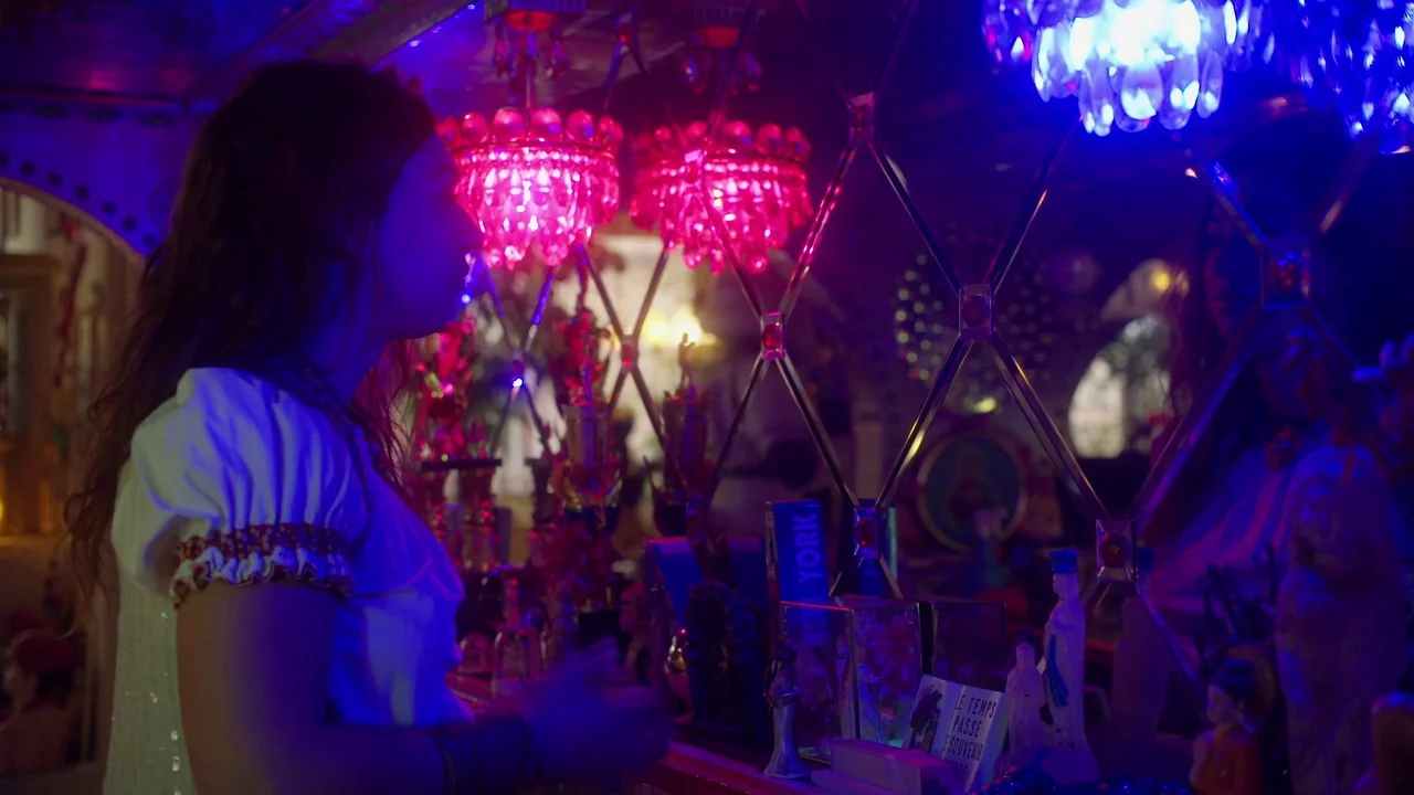 Orlando: My Political Biography (2023) movie still: medium — A dark retail space contains shelves filled with small figurines and souvenir items. A…