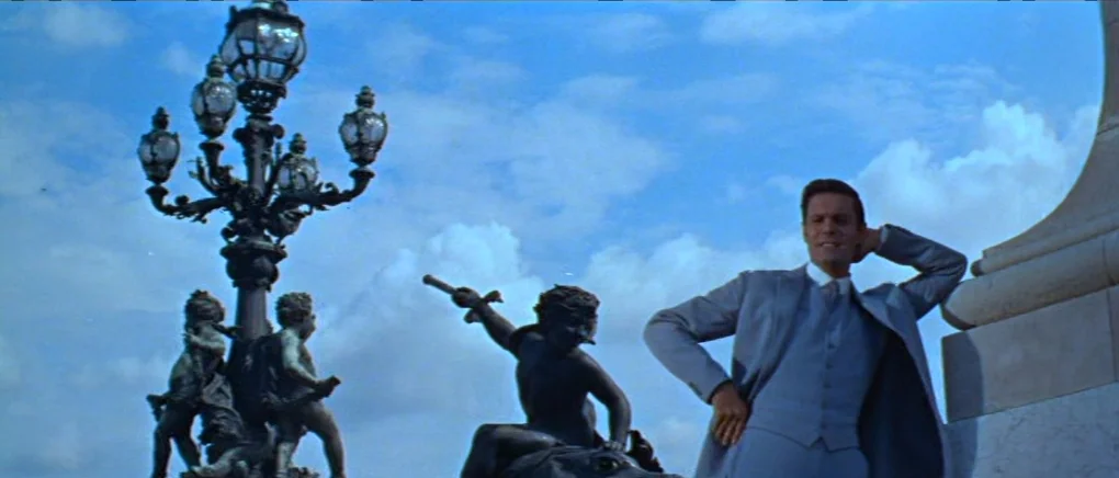 Gigi (1958) movie still: fullbody — A stone bridge parapet features ornate bronze sculptures of children and an elaborate lamp post. A man stands…
