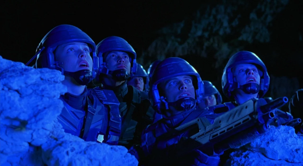 Starship Troopers (1997) movie still: medium — A group of soldiers in blue tactical armor hide behind a jagged rock formation. They gaze intensely…