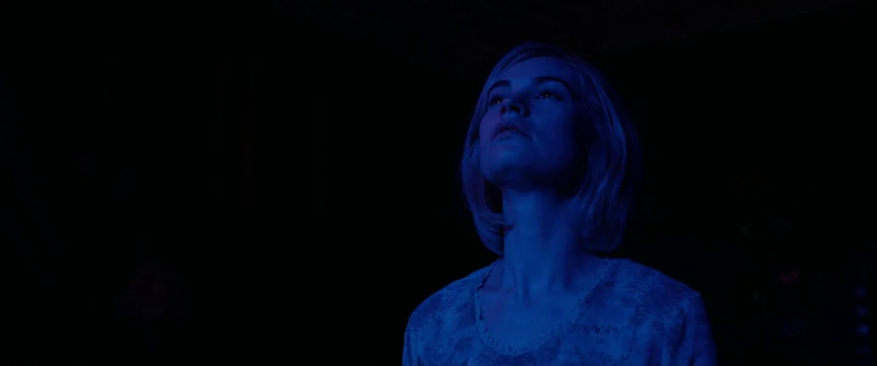 Rebecca movie still: medium — A dark indoor space with minimal visibility of the surroundings. A woman with blonde hair looks upward while…