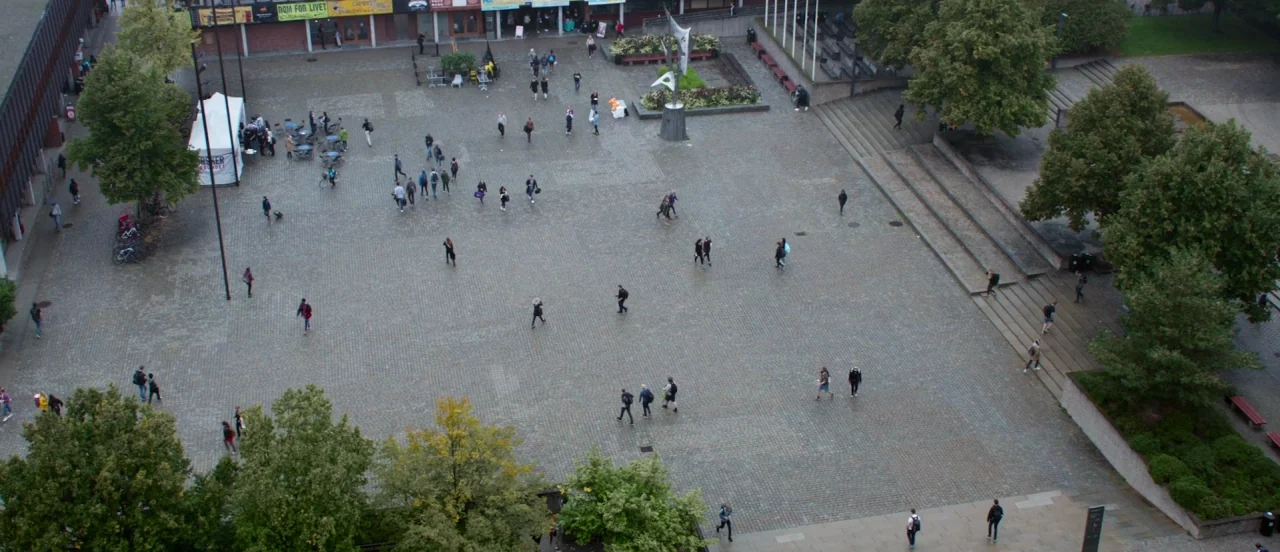 Thelma (2017) movie still: establishing — A large stone-paved urban plaza is surrounded by trees and modern brick buildings. Numerous individuals…