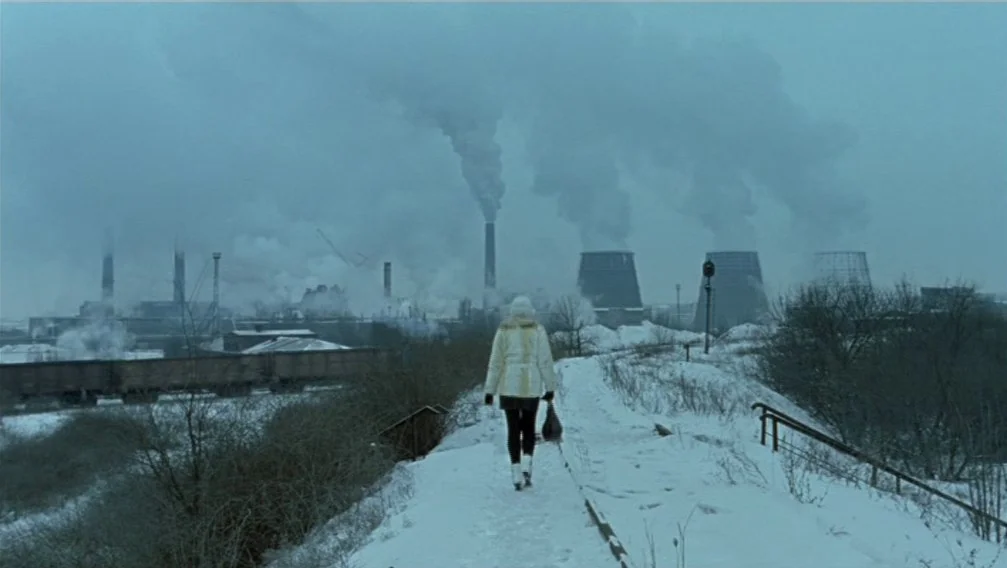 Import Export (2007) movie still: fullbody — A snowy, desolate industrial landscape with smoke-emitting cooling towers and train cars in background.…