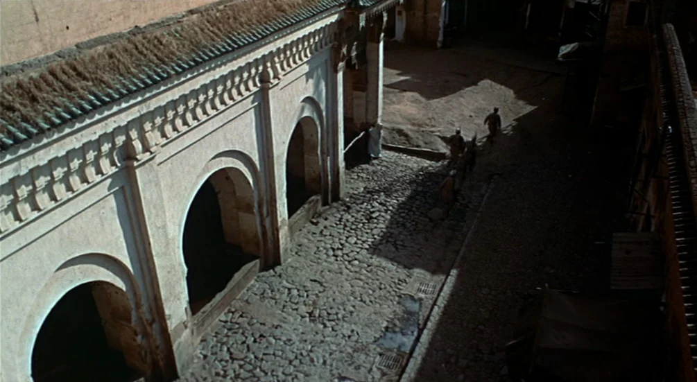 The Man Who Knew Too Much (1956) movie still: establishing — An exterior courtyard with arched walls made of pale stone and a cobblestone ground.…