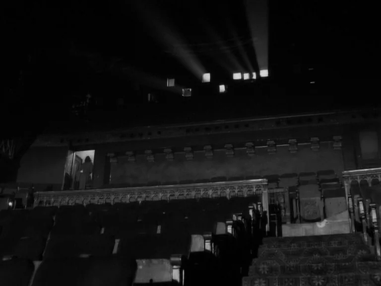The Good German movie still: wide — An empty movie theater auditorium features rows of seats facing toward a screen. Sharp beams of light emanate…