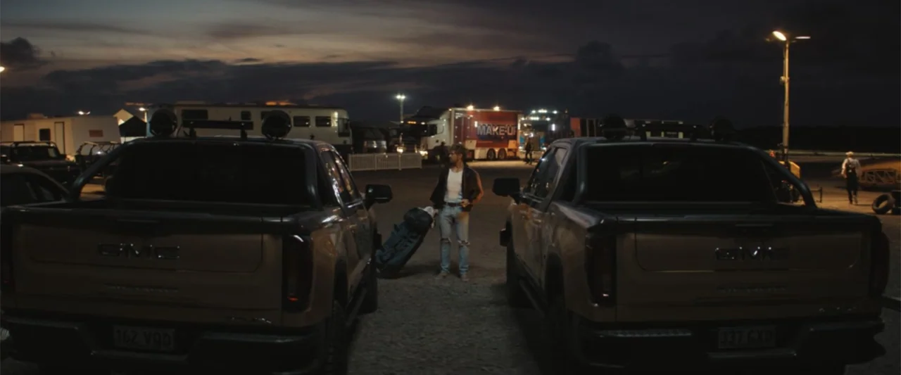 The Fall Guy (2024) movie still: wide — A gravel parking lot at night contains two large GMC pickup trucks parked symmetrically. A lone man stands…