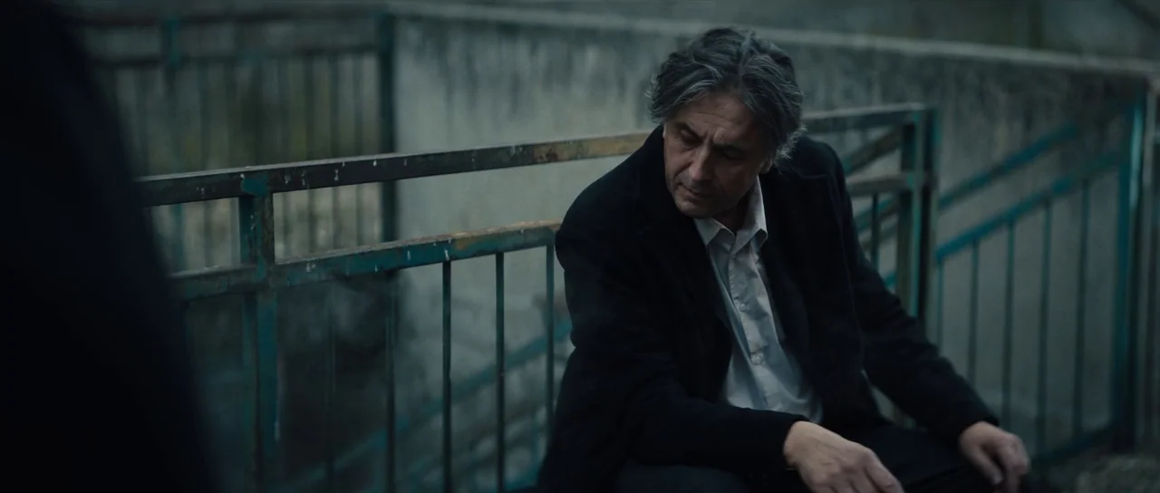 Hellraiser (2022) movie still: medium — A man sits on concrete stairs in an outdoor area with a rusted metal railing. He gazes downward towards an…