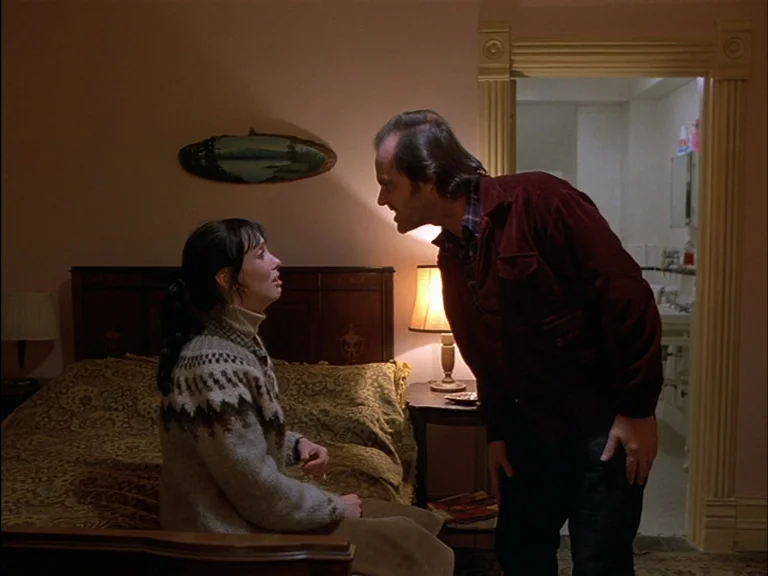 The Shining (1980) movie still: medium — A dimly lit room with floral patterned bedspreads and a lamp on a nightstand. A man in a dark jacket leans…