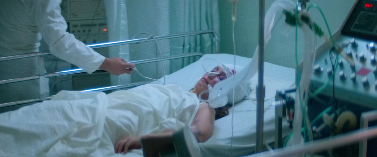 Vigilante (1982) movie still: medium — A medical setting with a patient lying in a bed surrounded by equipment. A person reaches to adjust the…