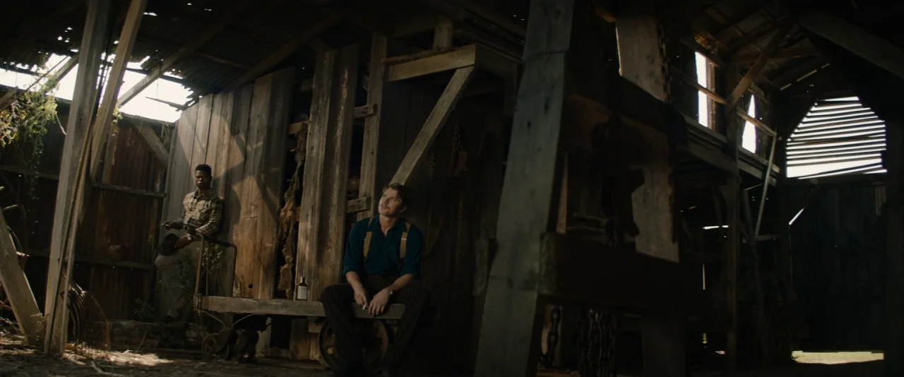 Mudbound (2017) movie still: wide — A rustic barn interior features weathered wooden beams, rusted metal walls, and scattered farm tools. Two men…