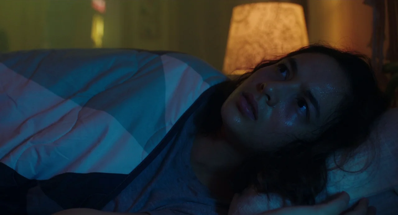 May The Devil Take You (2018) movie still: closeup — A dimly lit room features a person lying under blankets at night. The subject lies on their…