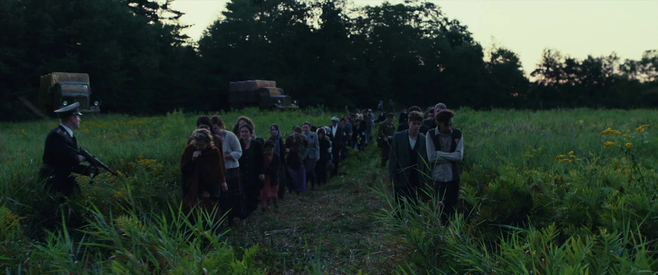 The Man Who Killed Hitler and Then the Bigfoot (2018) movie still: wide — A long line of civilians walks through a grassy field under a darkening…