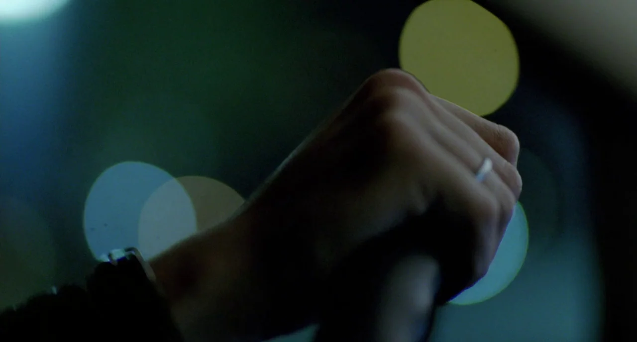 Tom at the Farm (2013) movie still: closeup — A close-up of a person's hands gripping a steering wheel inside a dark vehicle. Blurred urban city…
