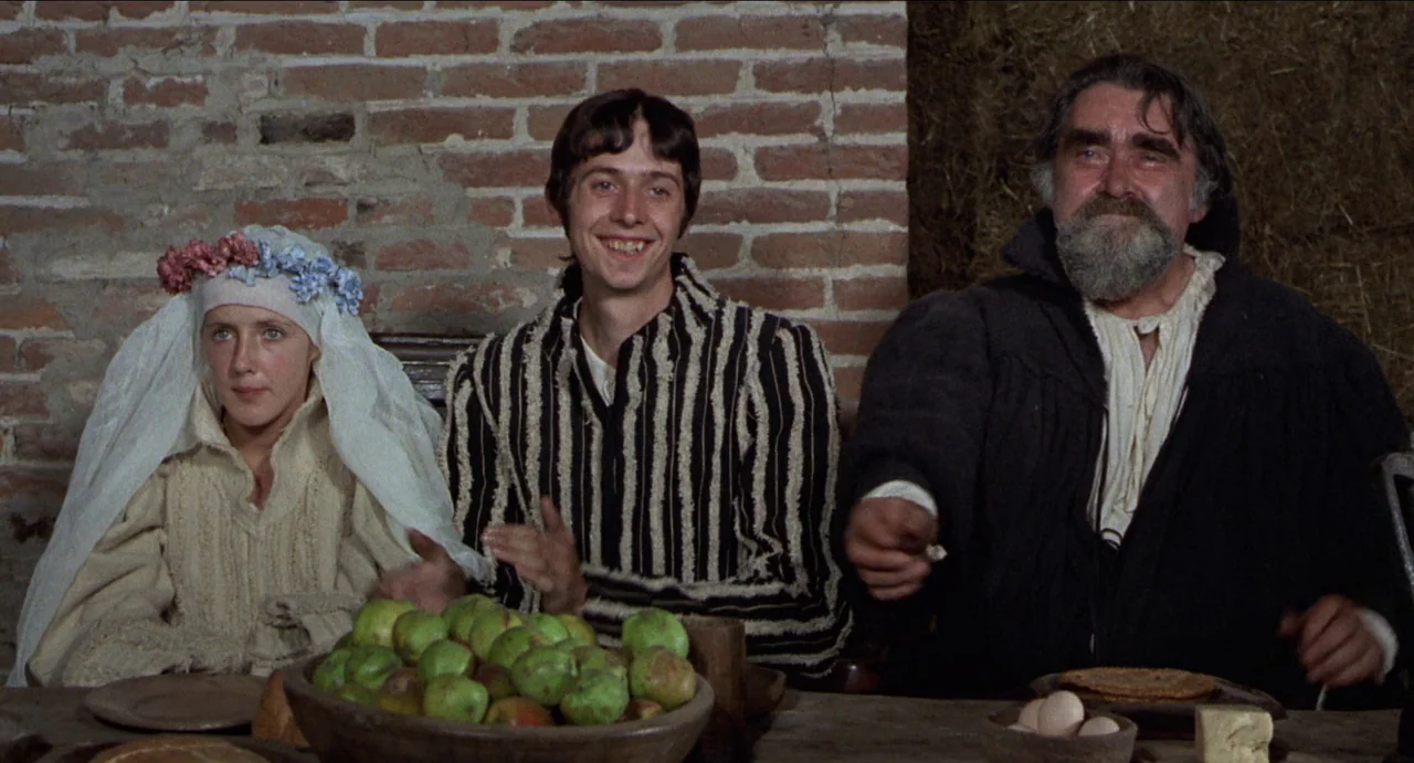 The Canterbury Tales (1972) movie still: medium — A rustic room featuring a brick wall and straw backing hosts a formal meal. Three people sit at a…