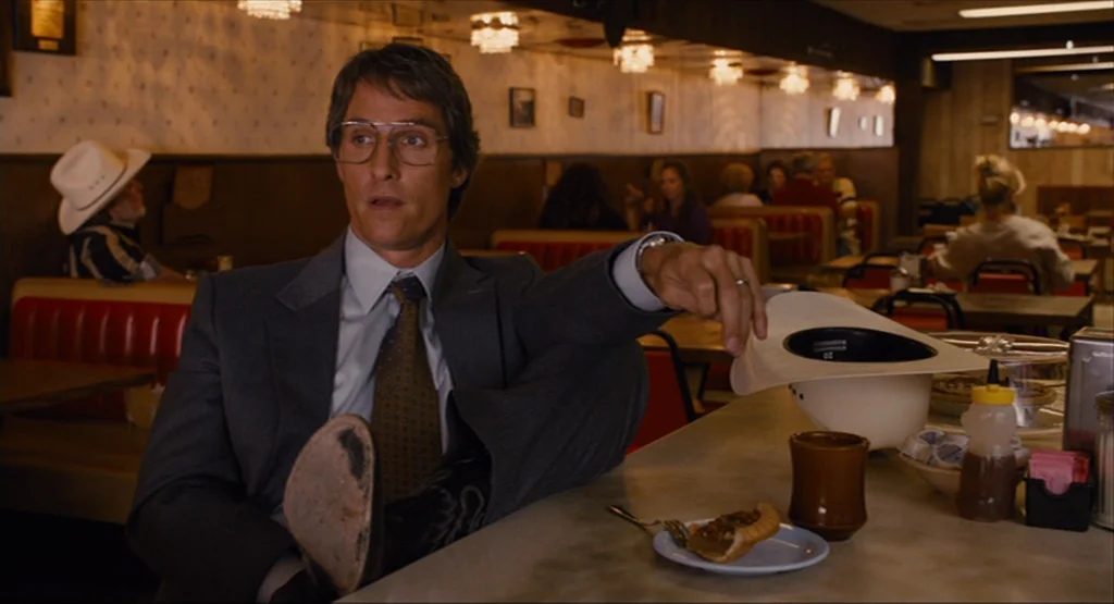 Bernie (2011) movie still: medium — A classic 1980s-style diner interior with red vinyl booths and wooden wall paneling. A man in a suit sits at the…