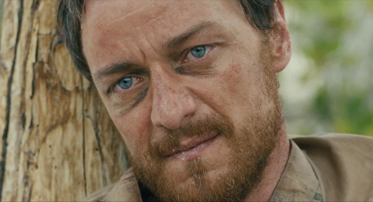 Submergence movie still: closeup — A man stands outdoors pressed against the rough bark of a textured tree trunk. He stares directly at the viewer…