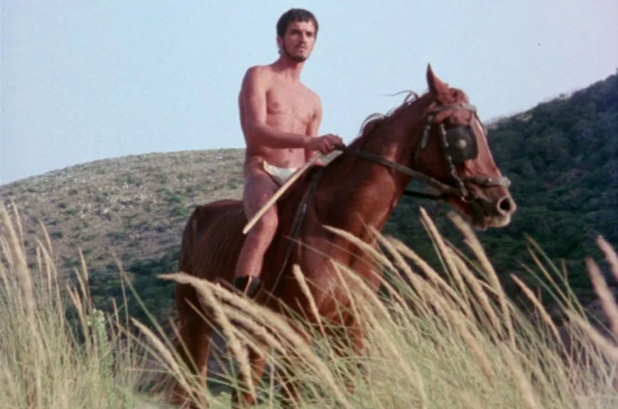 Sebastiane (1976) movie still: fullbody — A man rides a chestnut horse through a grassy, sun-drenched hillside landscape. The rider is shirtless and…