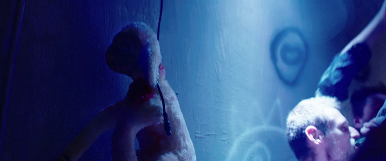 Raw (2016) movie still: medium — A dark, cramped basement with blue lighting and graffiti on the rough plaster wall. A man stands holding a worn…