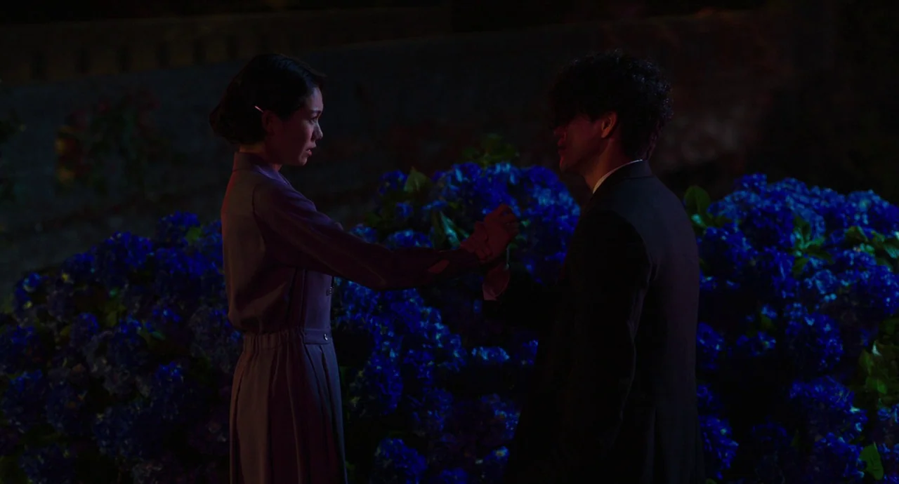 No Longer Human (2019) movie still: medium — A man and woman stand close together in front of a dense blue flowering bush. The woman holds the man's…