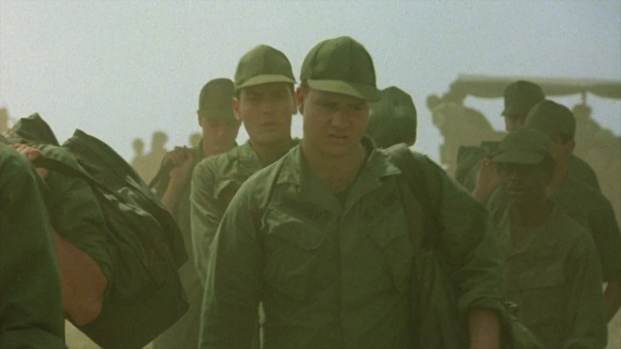 Platoon (1986) movie still: medium — A dusty, sunlit outdoor area shows a group of soldiers moving together. They wear matching green military…