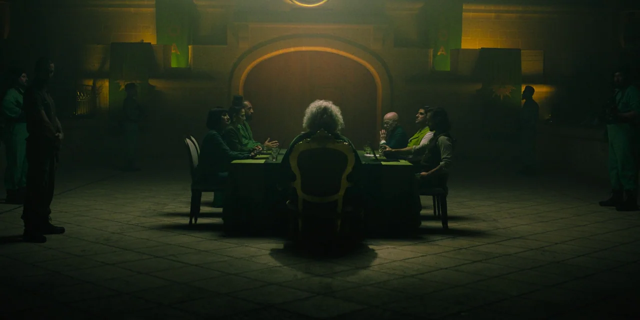 Sniper: G.R.I.T. (2023) movie still: wide — A stone-floored chamber features a large conference table illuminated by an overhead yellow light. A…