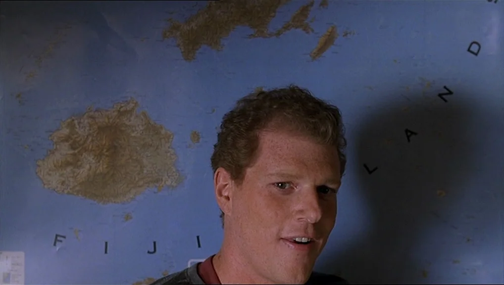 The Truman Show movie still: closeup — A room features a large, detailed wall map of the Fiji Islands. A man stands directly in front of the map…