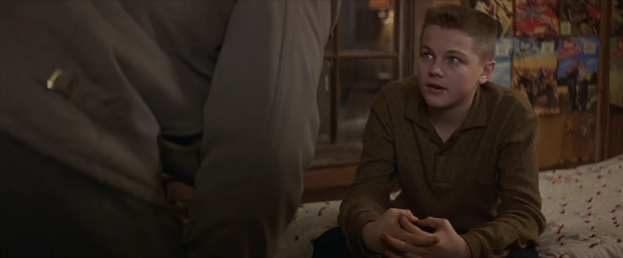 This Boy's Life (1993) movie still: medium — A 1950s bedroom features comic books tacked to the wall near a bed. A teenage boy sits on the edge of…