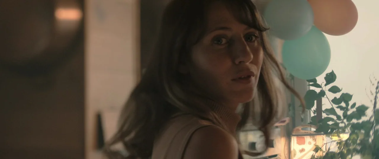 Oxygen (2021) movie still: closeup — A bright living room decorated with pastel balloons near a window with indoor plants. A woman turns her head to…