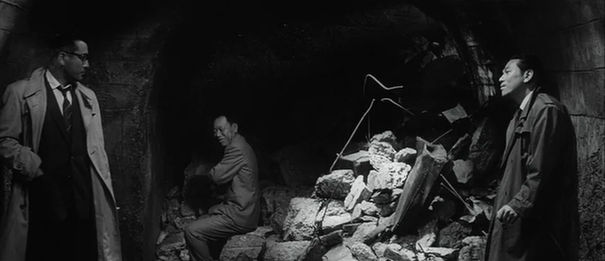 The Bad Sleep Well (1960) movie still: wide — A cavernous, dark tunnel contains a large pile of broken masonry and stone debris. Three men in formal…