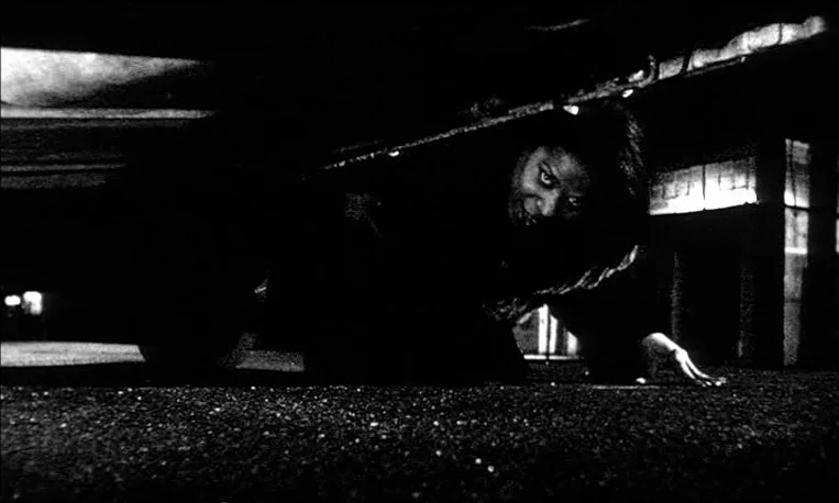 Pi (1998) movie still: medium — A dark, textured asphalt surface sits beneath a large vehicle chassis or low structure. A woman crawls through the…