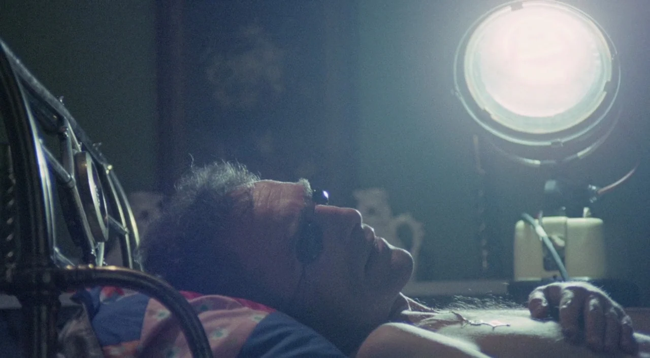 The Iguana with the Tongue of Fire (1971) movie still: medium — A dark, dimly lit space features a man resting on a bed under cool lighting. The man…