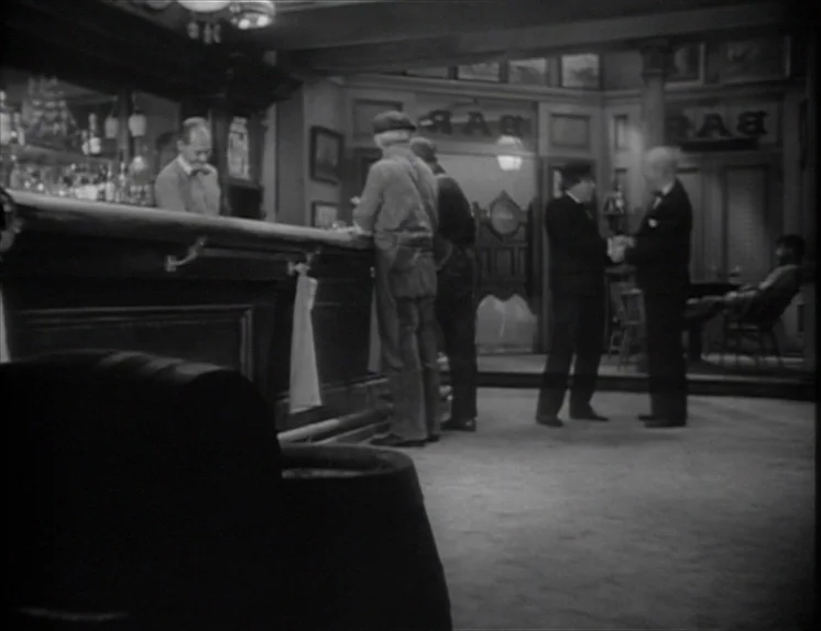 Anna Christie (1930) movie still: wide — A dimly lit 1940s pub interior with wooden bar, dark fixtures, and paneled walls. Several men stand at the…