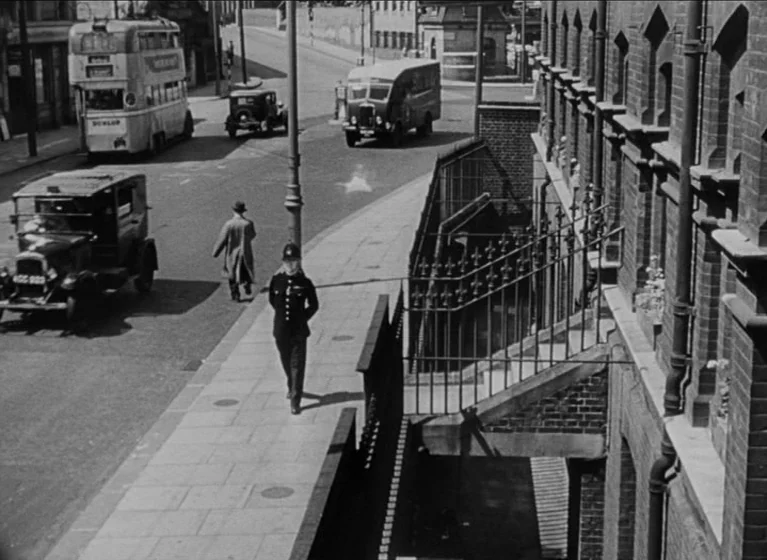 The Blue Lamp (1950) movie still: wide — An urban sidewalk runs alongside a tall brick building with an iron railed staircase. A British police…