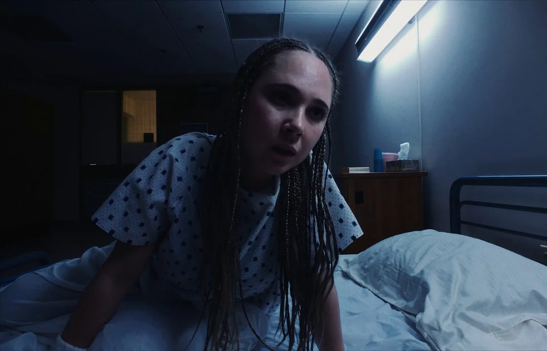 Unsane (2018) movie still: medium — A dark hospital room features a single bed and a small wooden nightstand. A woman in a hospital gown leans over…