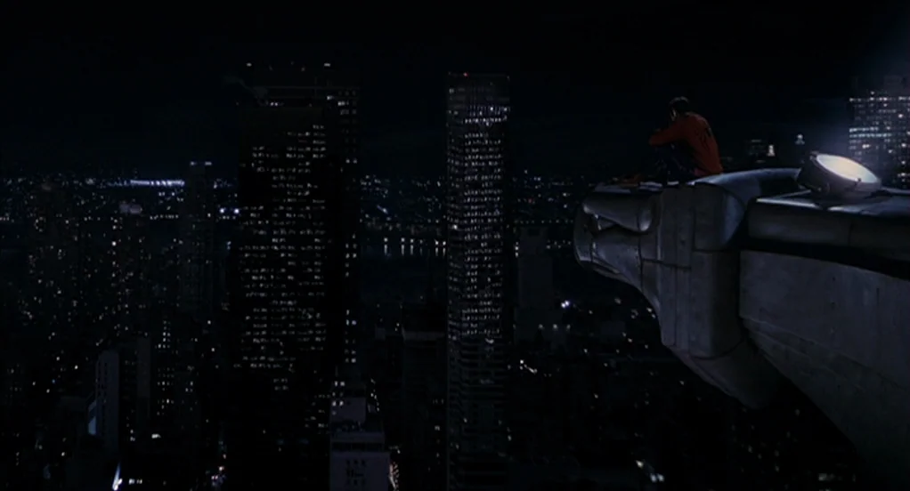 Spider-Man (2002) movie still: wide — A high-angle view looks over a dark city at night from a ledge. A person sits perched on a large stone…