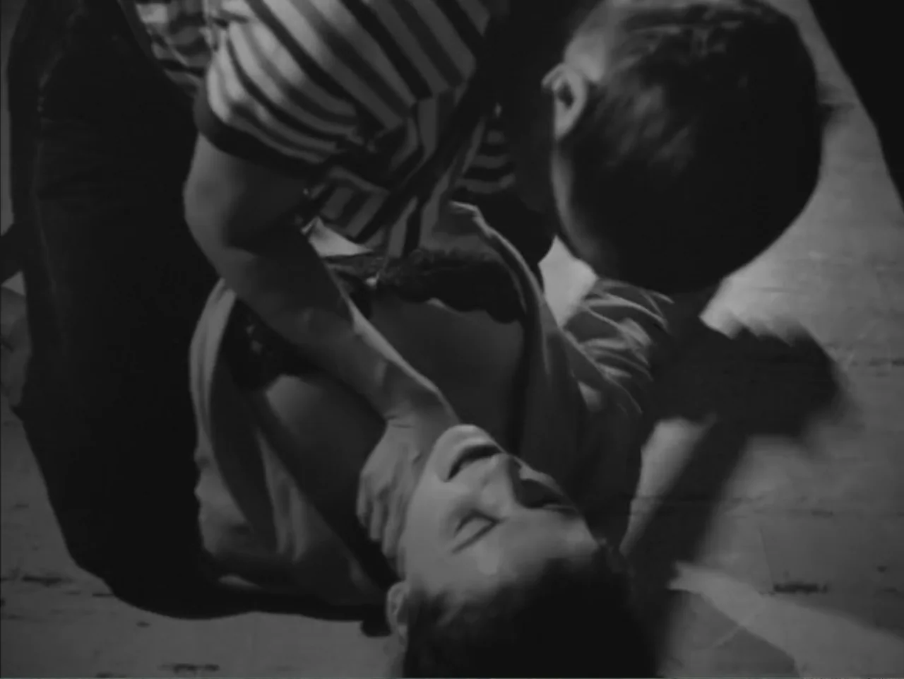 Chained Girls (1965) movie still: medium — Two individuals occupy a dark space on a smooth floor surface. A man leans over a woman, holding her neck…