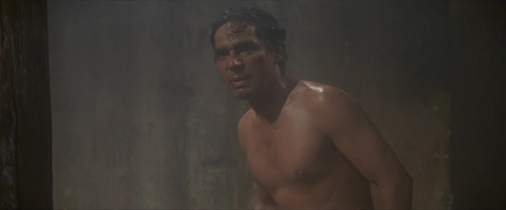 The Wild Bunch (1969) movie still: medium — A dark, dimly lit stone enclosure with textured walls. A shirtless man stares off-screen with sweat…