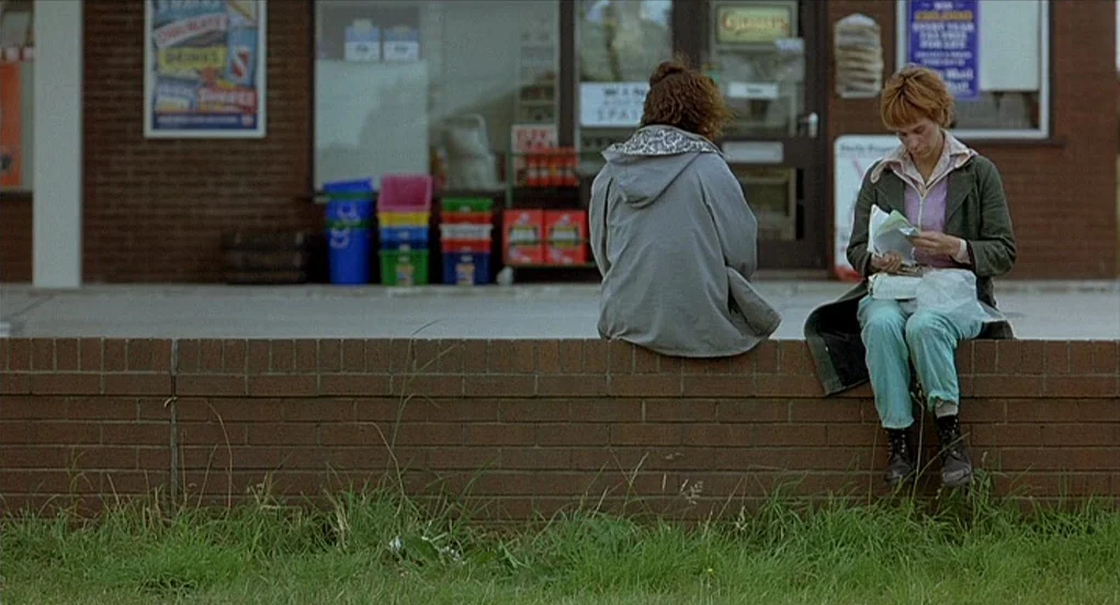 Butterfly Kiss (1995) movie still: fullbody — A low brick wall stands before a commercial storefront with visible advertising posters. Two young…