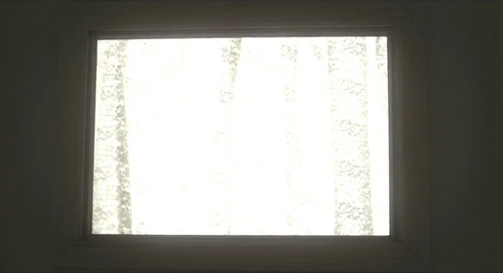 The Future (2011) movie still: closeup — A rectangular window frame sits against a darkened wall in an interior space. Beyond the glass, the faint…