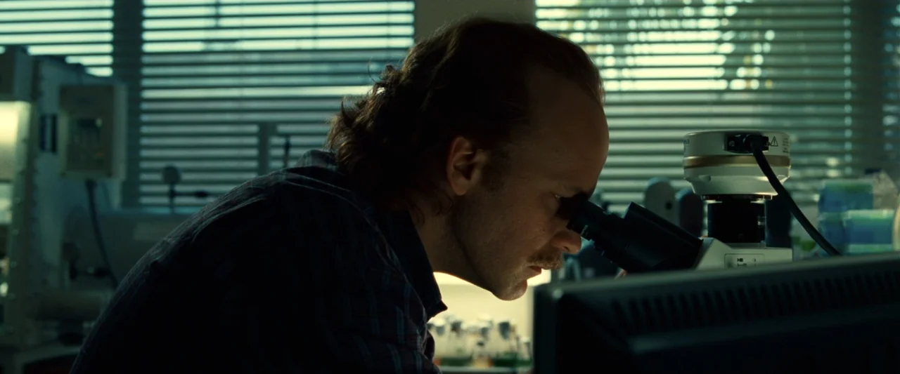 Green Lantern (2011) movie still: medium — A laboratory setting features a desk with a microscope positioned in front of closed blinds. A man in a…