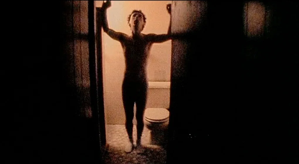 Julien Donkey Boy (1999) movie still: fullbody — A dimly lit room viewed through a dark doorway with a toilet visible inside. A nude man stands…