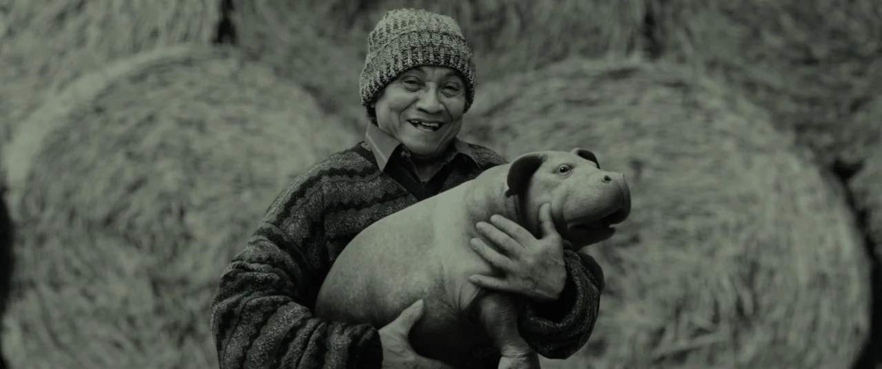 Okja (2017) movie still: medium — The background consists of several large cylindrical hay bales stacked together. An older man wearing a knit…