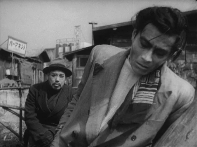 Drunken Angel (1948) movie still: medium — A cluttered outdoor area features low-lying wooden structures and a sign with Japanese text. One man in a…