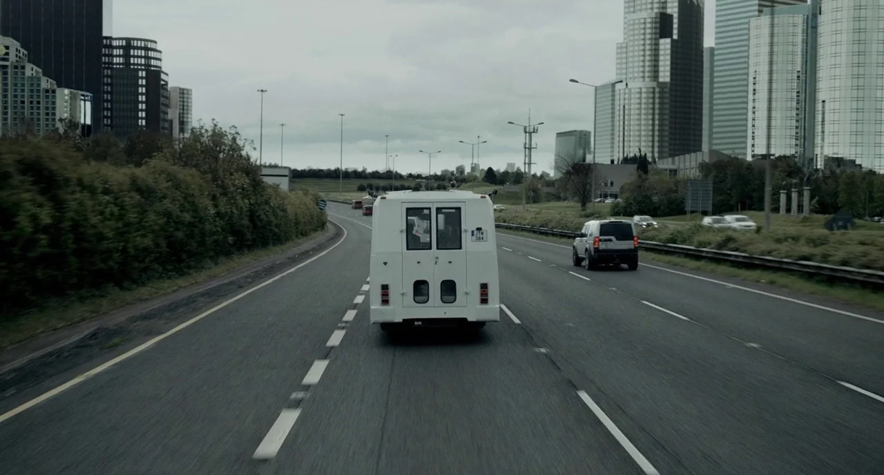 The Lobster (2015) movie still: wide — A multi-lane highway stretches toward a distant modern city skyline under an overcast sky. A white van moves…