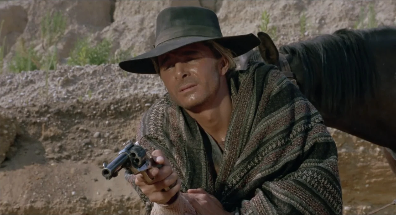 A Stranger in Town (1943) movie still: medium — An arid landscape features sandy ground and a sparse hillside under bright daylight. A man in a…