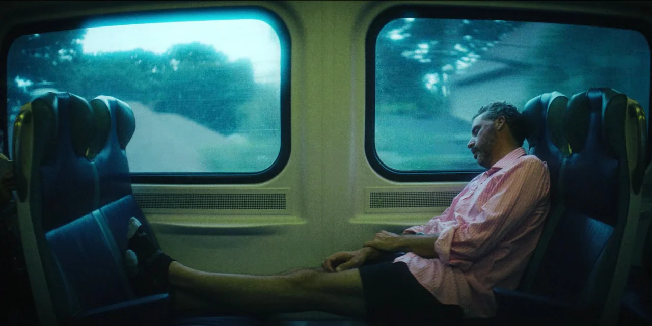 Spermworld (2024) movie still: medium — A man sleeps across two blue train seats in a dimly lit passenger carriage. The windows show a blurred green…